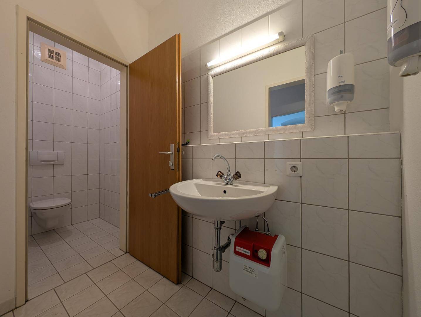 Funktionales Badezimmer mit Waschbecken, Spiegel und gefliesten Wänden, inklusive separatem WC-Bereich.