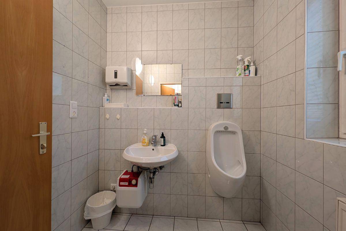 Gefliestes Badezimmer mit Urinal, Waschbecken und Fenster, ausgestattet mit hellen Wand- und Bodenfliesen.