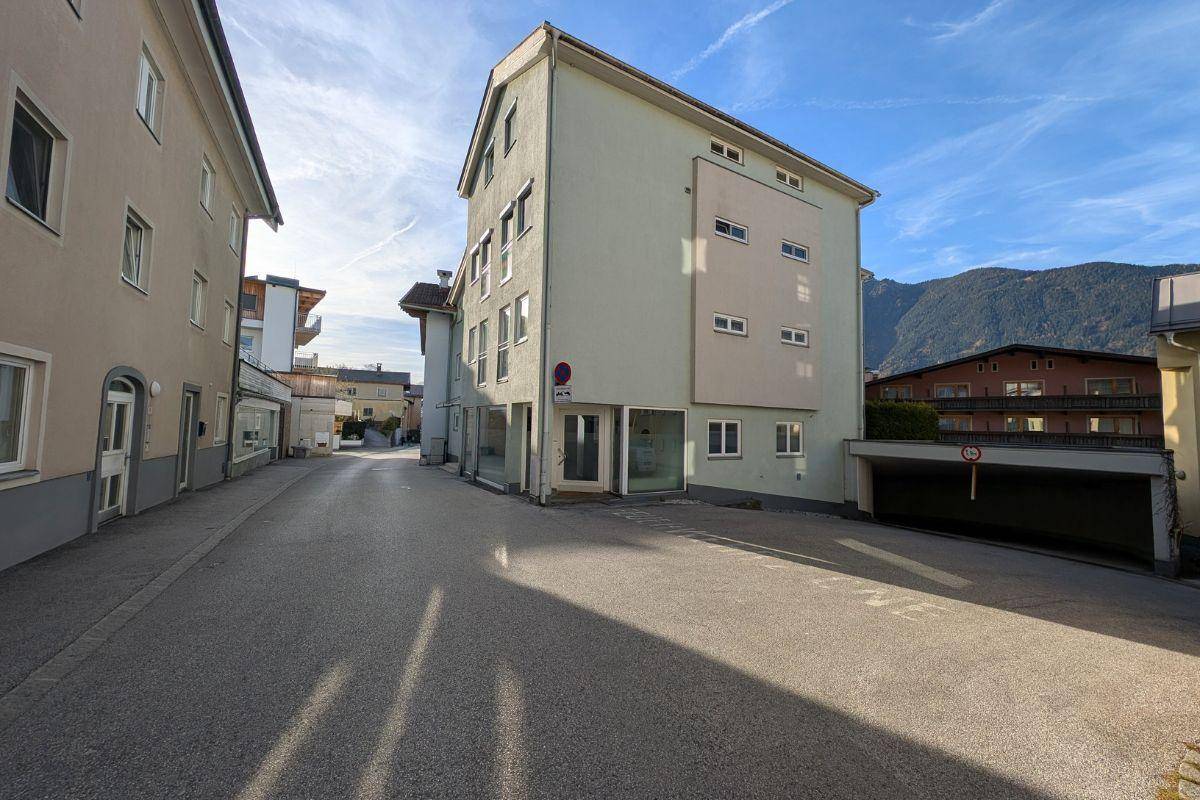Mehrstöckiges Gebäude mit hellgrüner Fassade an einer Straße, mit Tiefgarageneinfahrt und Bergblick im Hintergrund.