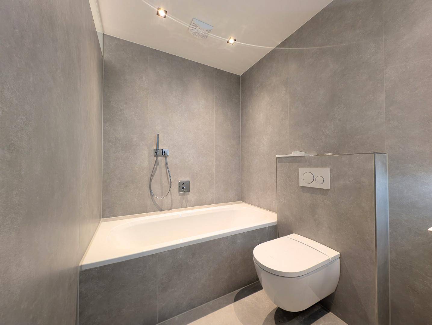 Badezimmer mit integrierter Badewanne, Wand-WC und modernen grauen Fliesen.