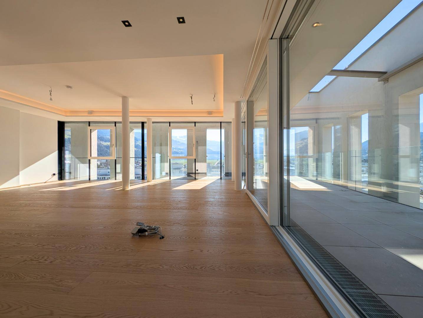 Heller, leerer Wohnbereich mit großen Fensterfronten, Holzboden und Zugang zur Terrasse.