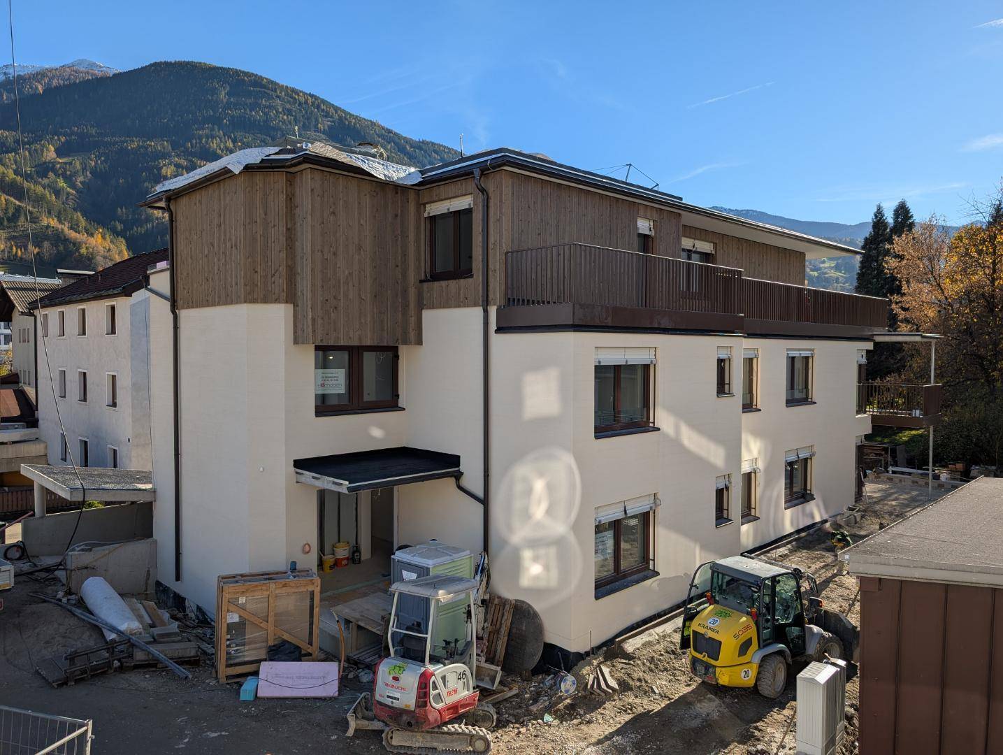 Ansicht des Neubaus von der Rückseite mit Baugeräten und der umliegenden Berglandschaft.