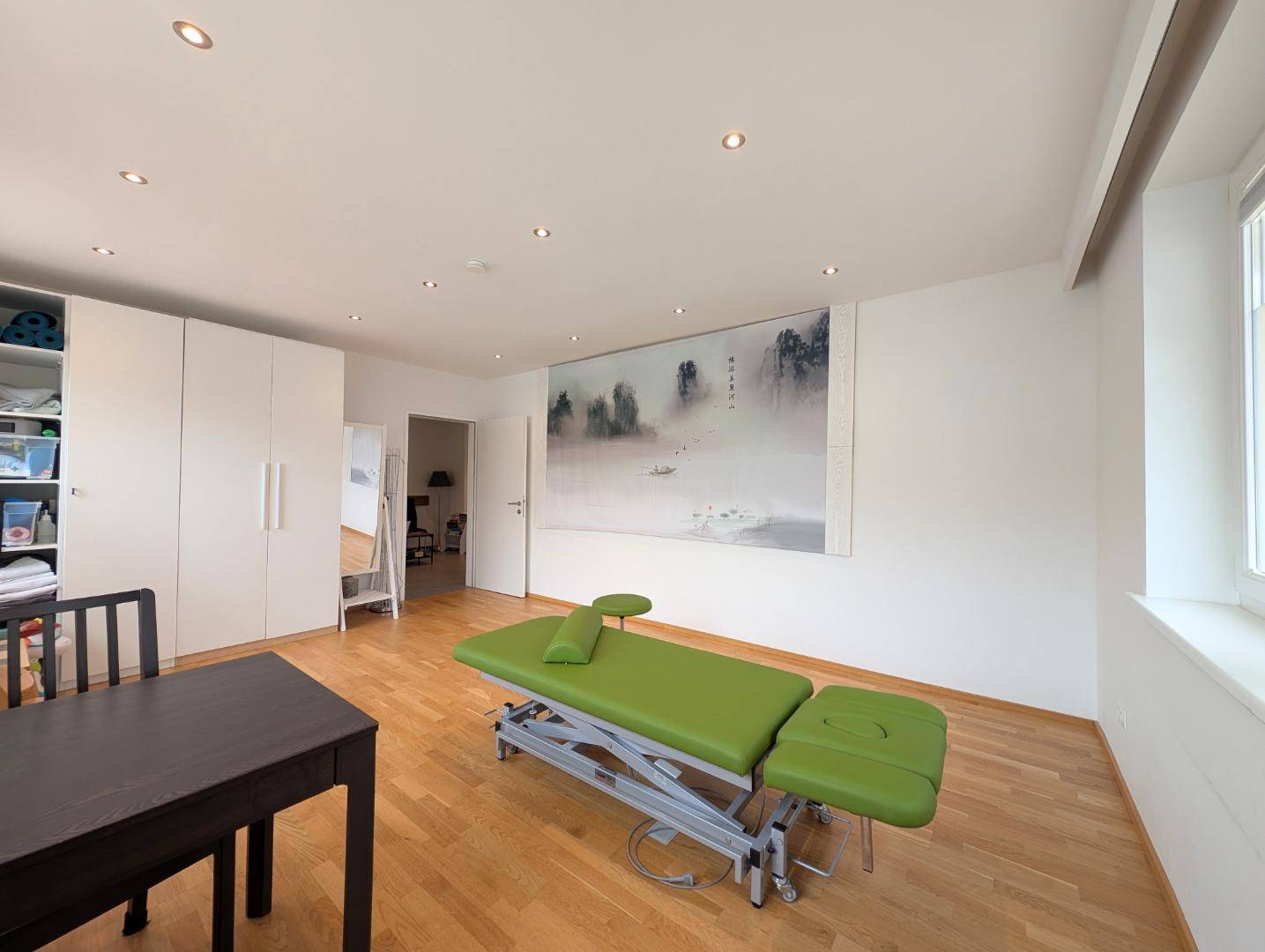 Therapieraum mit weißem Wandschrank, Behandlungsliege und einem großen Wandbild als Dekoration.