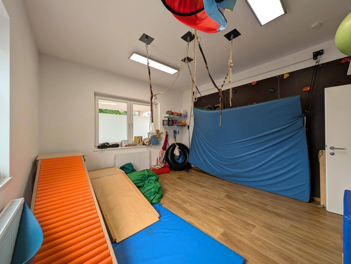 Therapieraum mit Holzboden, Kletterwand und verschiedenen Therapiegeräten.