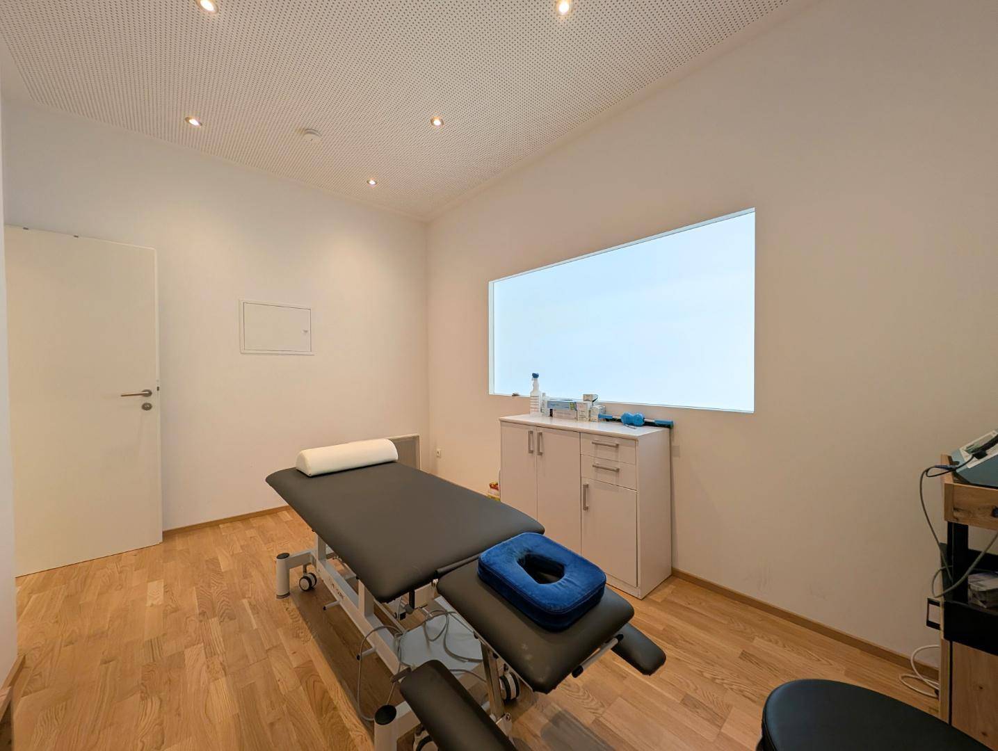 Therapieraum mit Holzboden, Behandlungsliege und einem großen Fenster.