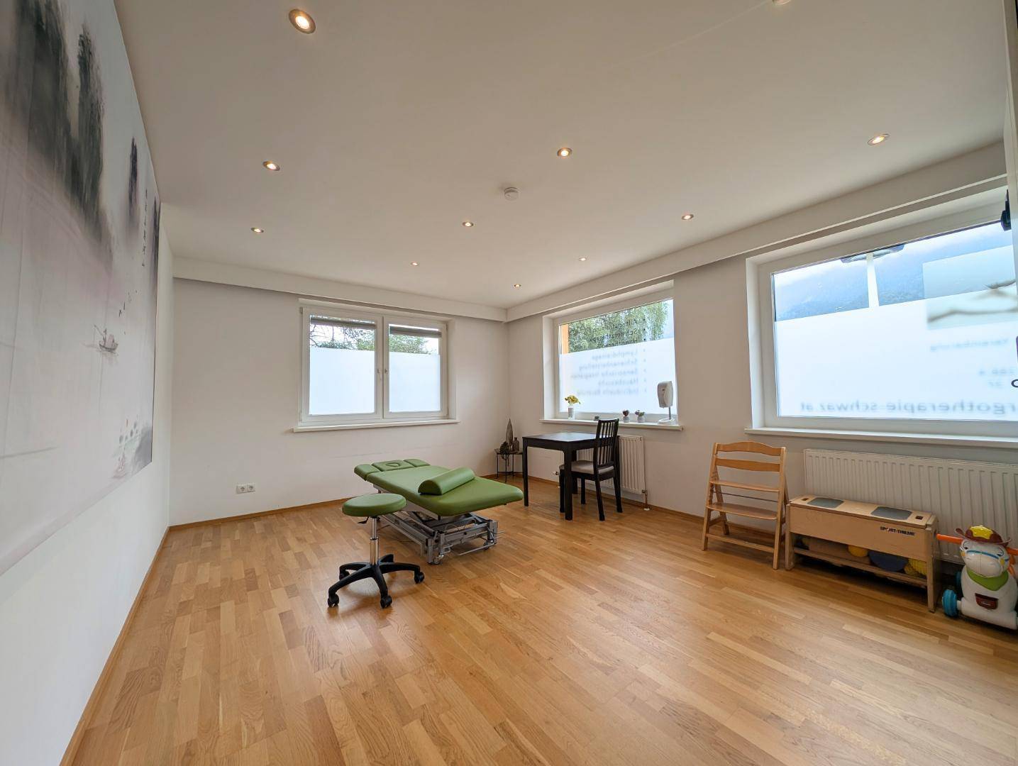 Geräumiger Therapieraum mit Holzboden, Behandlungsliege und großen Fenstern für viel Tageslicht.