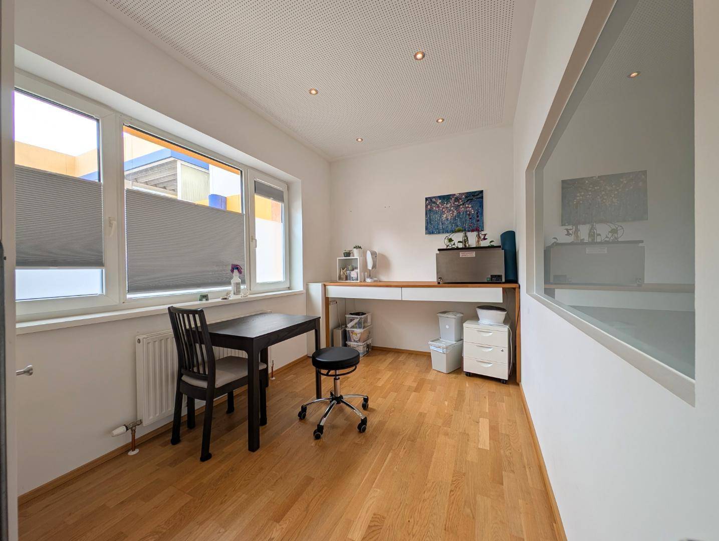 Helles Büro mit Holzboden, Schreibtisch und großen Fenstern mit Jalousien.