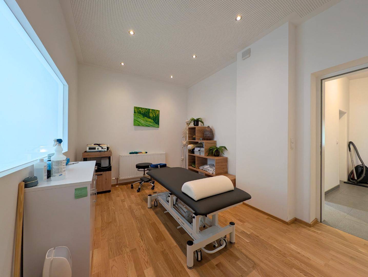 Therapieraum mit Holzboden, Behandlungsliege und Regalen für Materialien.