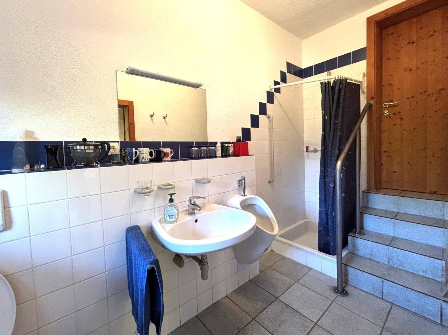 Badezimmer mit weißen Fliesen, Dusche, Urinal und Waschbecken.