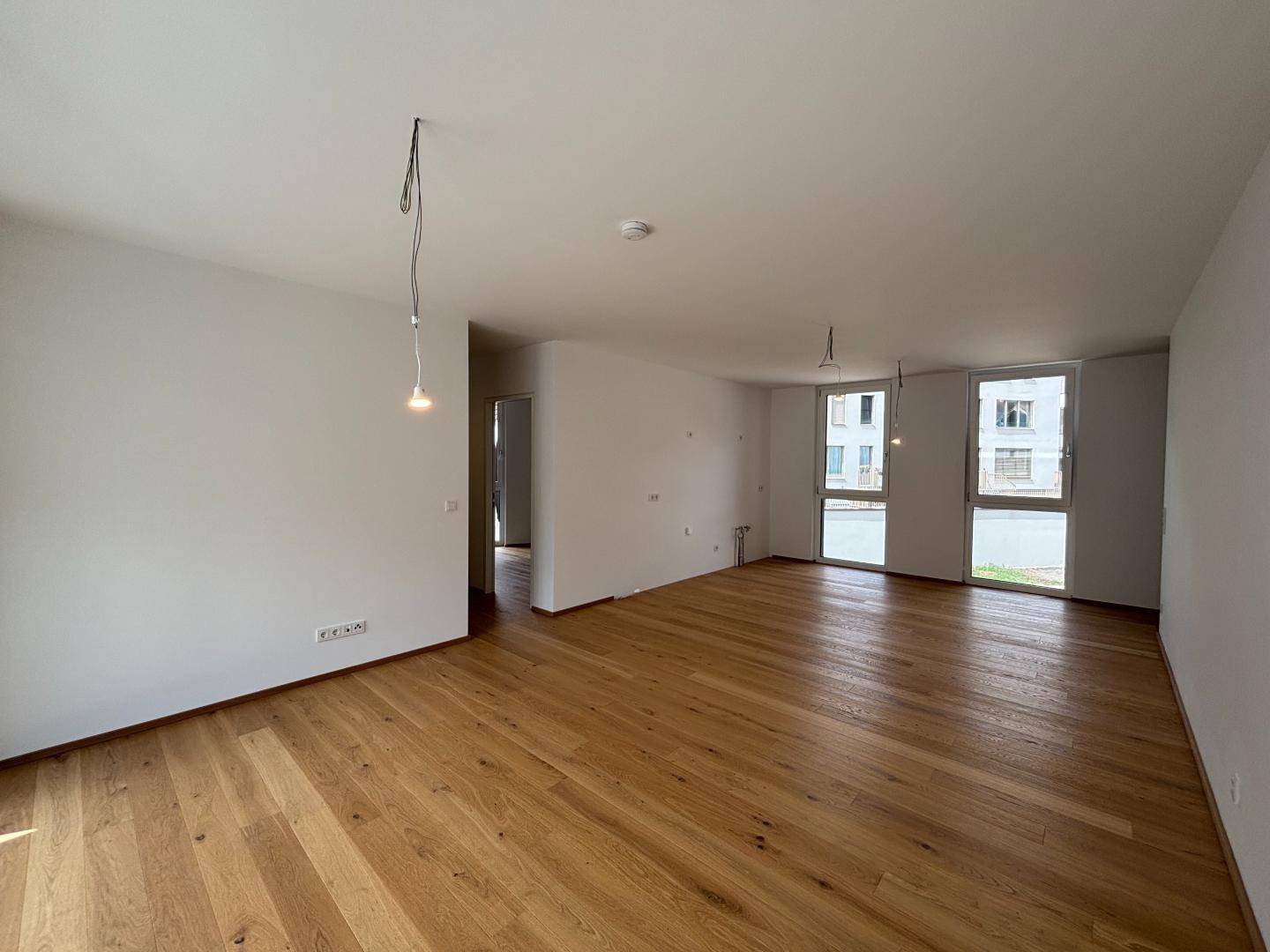 Geräumiger, unmöblierter Wohnbereich mit hellem Holzparkett und großen Fenstern.
