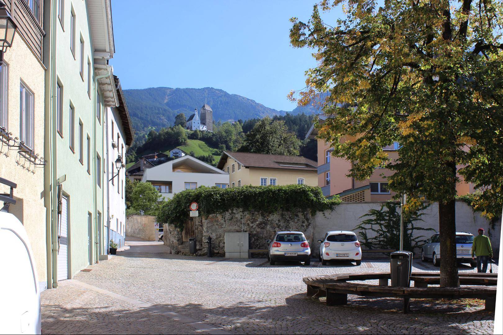 Blick auf eine ruhige Gasse in einer Bergstadt mit traditionellen Gebäuden und einem modernen Neubau im Hintergrund.