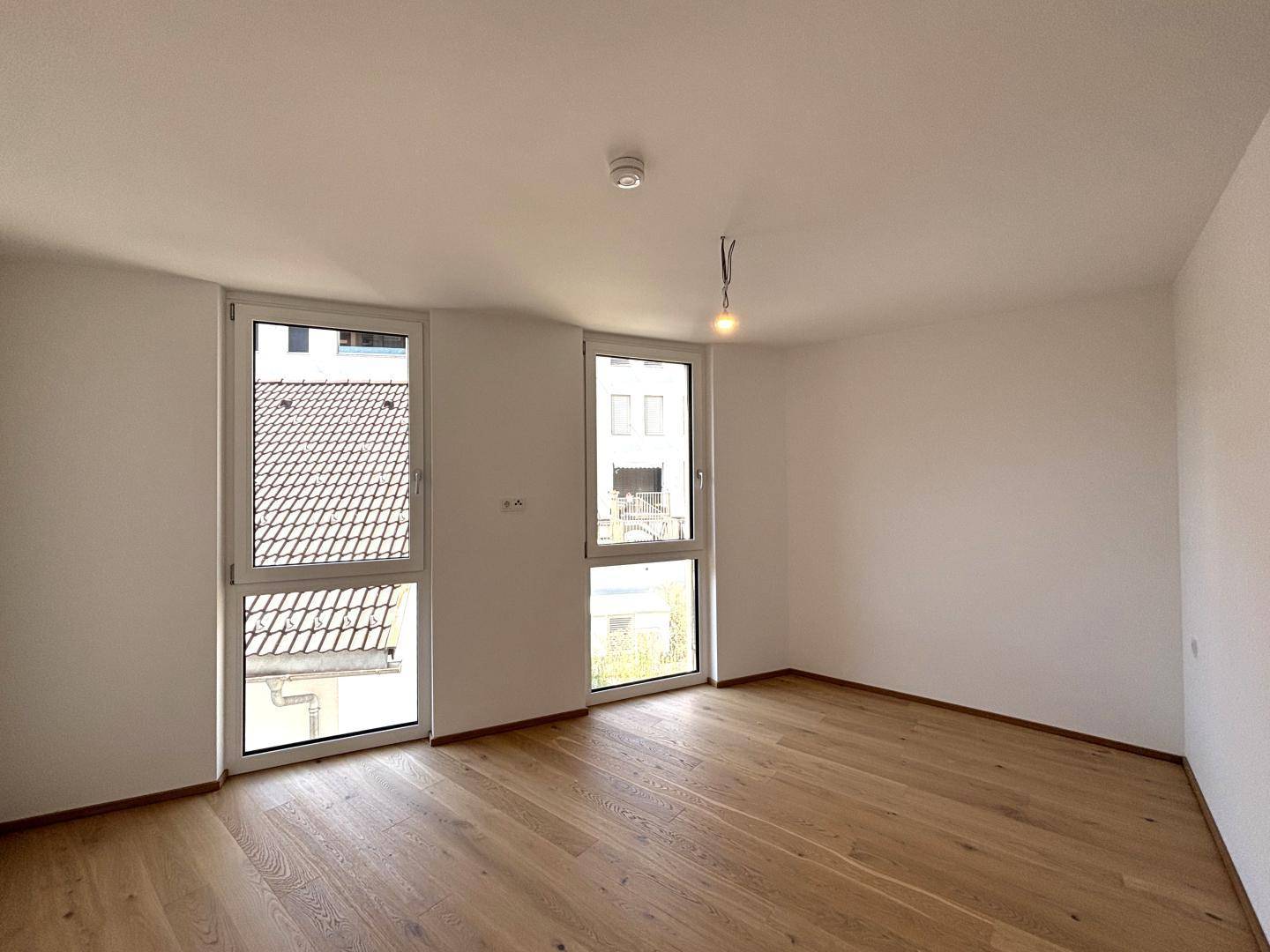 Heller Raum mit Holzboden und zwei hohen Fenstern, die viel Tageslicht hereinlassen.
