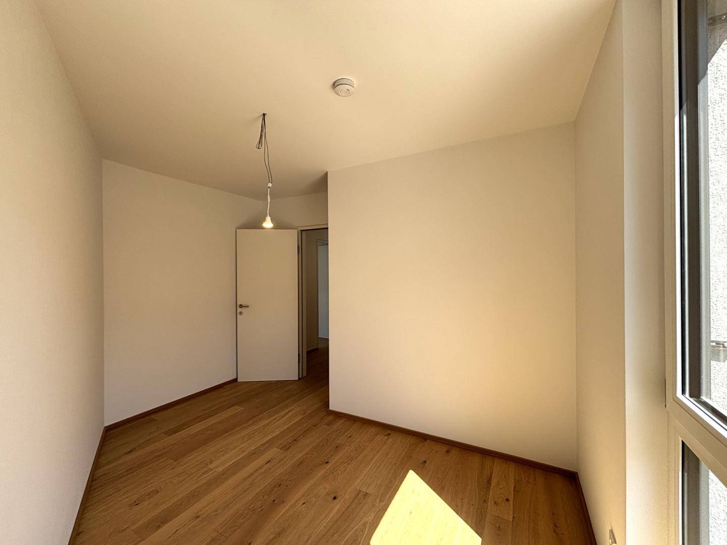 Leerer Raum mit Holzboden und einem hohen Fenster, das den Raum mit natürlichem Licht füllt.