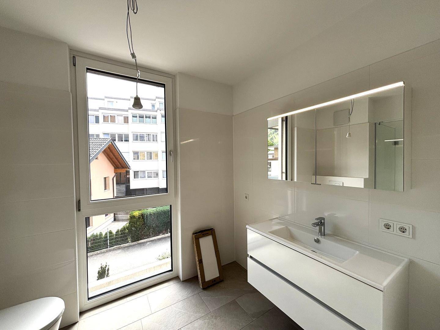 Modernes Badezimmer mit Waschtisch, beleuchtetem Spiegel und großem Fenster für Tageslicht.