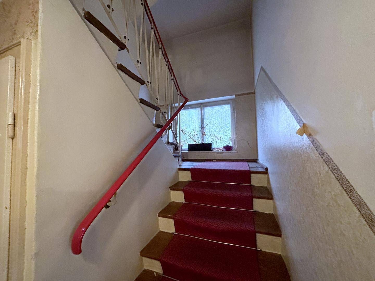 Treppenhaus mit rotem Teppich und Geländer, das zu einem hellen Fensterbereich führt.
