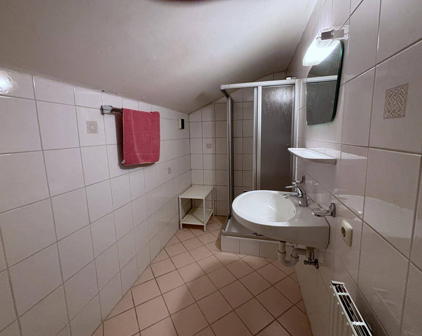 Gefliestes Badezimmer mit Duschkabine, Waschbecken und Heizkörper, unter einer Dachschräge.