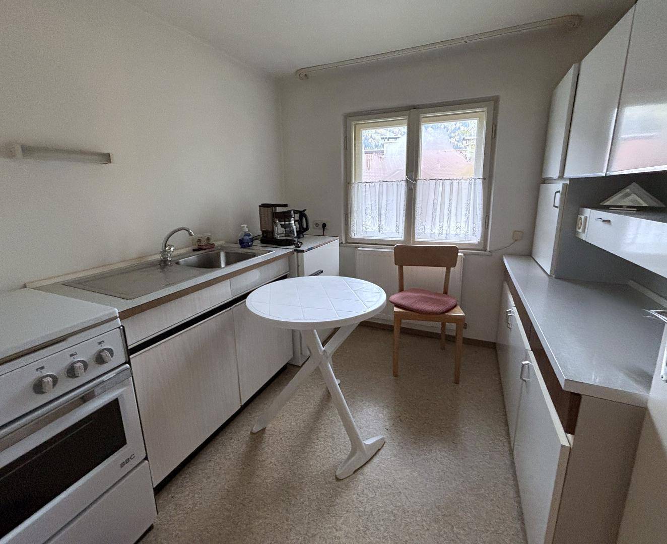 Kleine Küche mit weißen Schränken, Herd, Spüle und einem runden Tisch, gut beleuchtet durch ein Fenster.