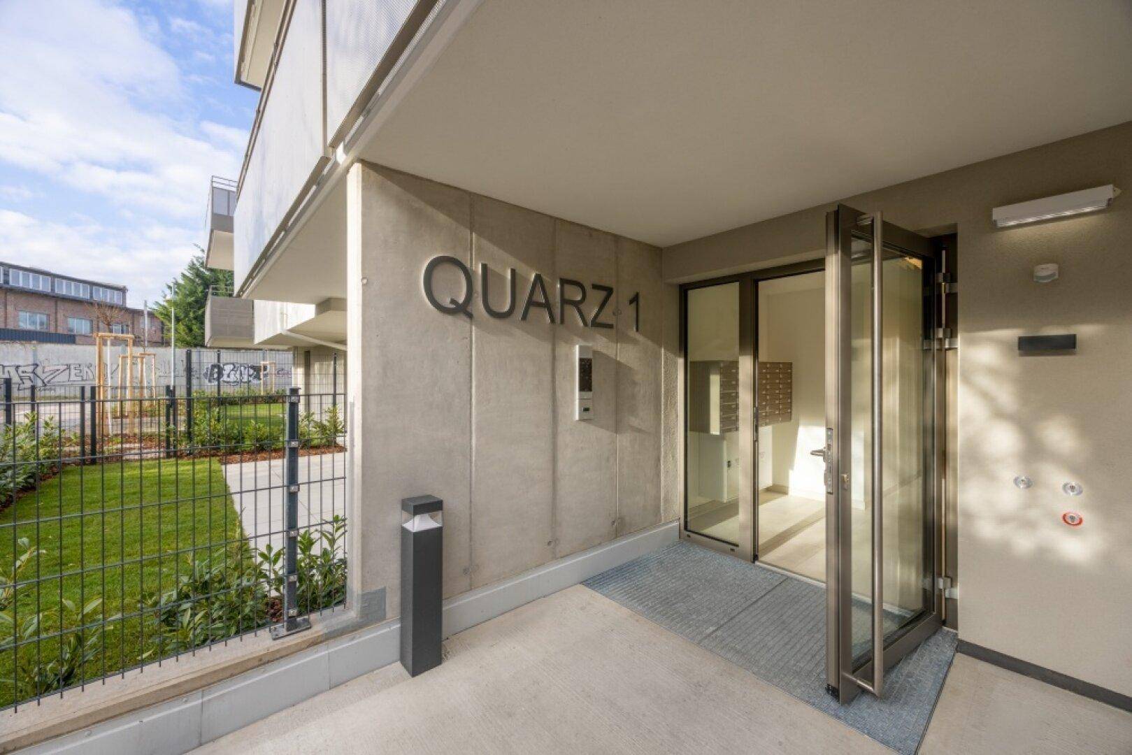 Eingangsbereich des modernen Gebäudes 'QUARZ 1' mit Rampe, Glastür und Namenszug.