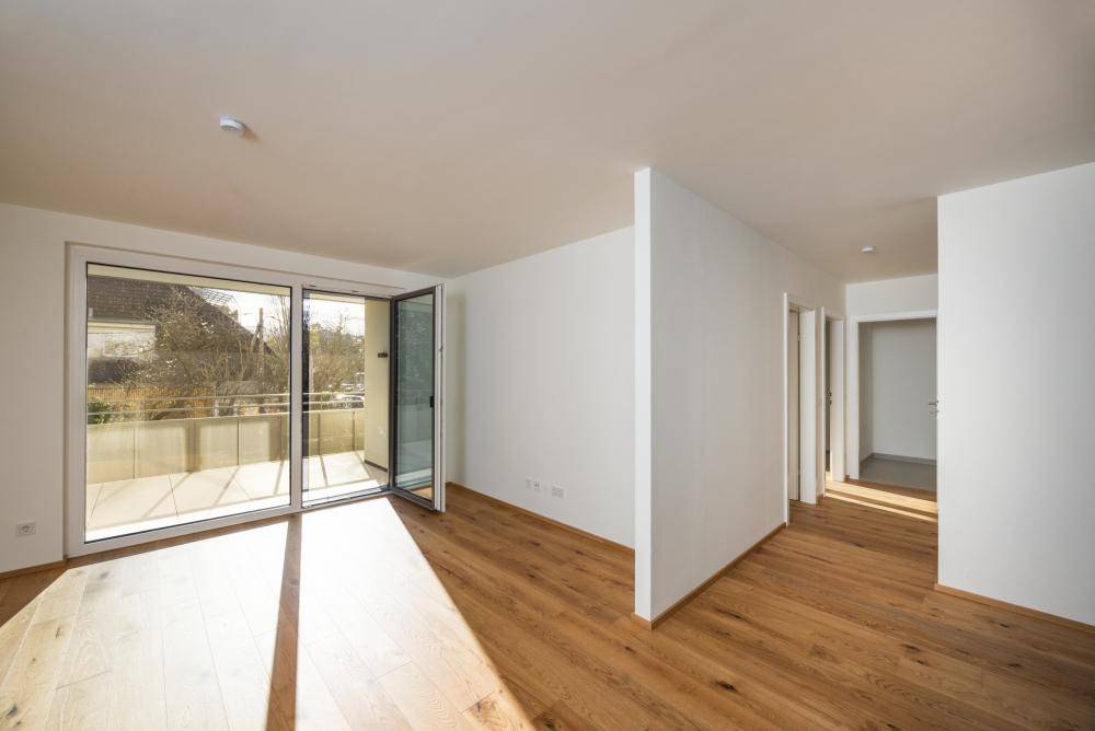 Lichtdurchflutetes Zimmer mit Holzfußboden und großem Schiebefenster, das zum Balkon führt.