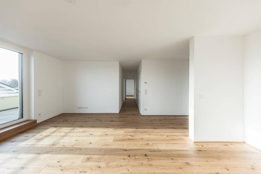 Helles, leeres Zimmer mit Holzboden und großen Fenstern, die viel Tageslicht hereinlassen.