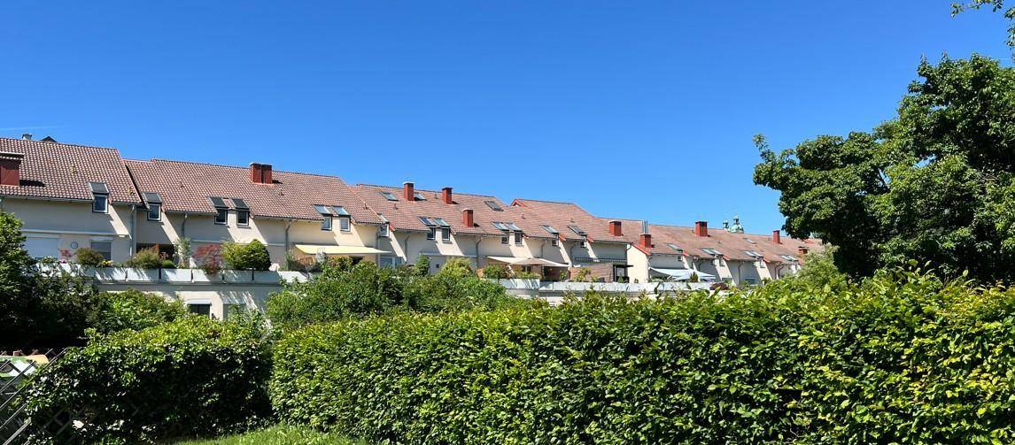 Reihe von Wohnhäusern mit roten Ziegeldächern und grünen Hecken im Vordergrund unter blauem Himmel.