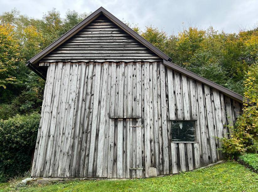 Rustikales Holzgebäude mit vertikalen Holzbrettern und einem kleinen Fenster, umgeben von Bäumen.