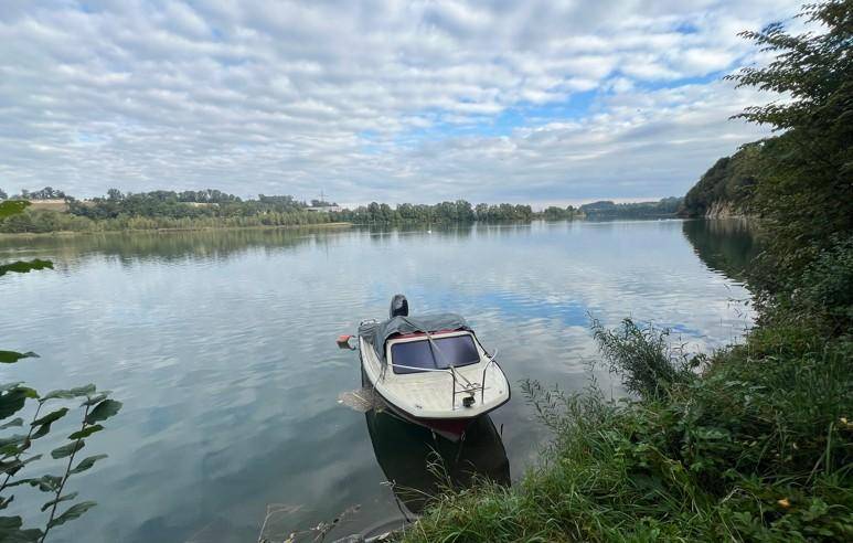 Ein kleines Boot schwimmt ruhig auf einem See unter einem bewölkten Himmel, umgeben von Ufervegetation.