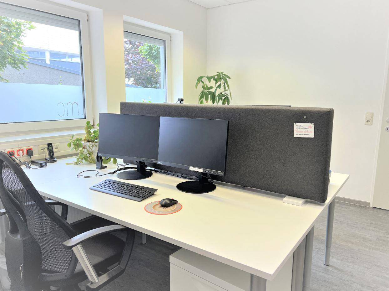 Moderner Büroarbeitsplatz mit zwei Monitoren, ergonomischem Stuhl und viel Tageslicht durch das Fenster.