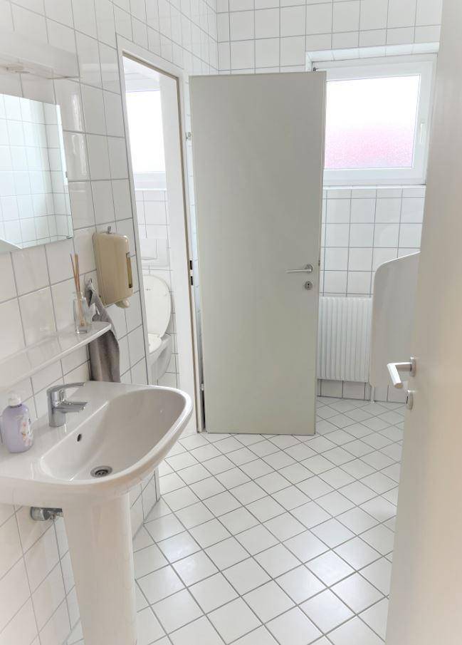 Helles Badezimmer mit weißen Fliesen, Waschbecken, Urinal und Fenster für natürliche Belüftung.