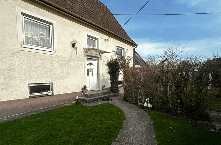 Eingangsbereich eines Hauses mit gepflastertem Weg, kleiner Rasenfläche und Vordach.