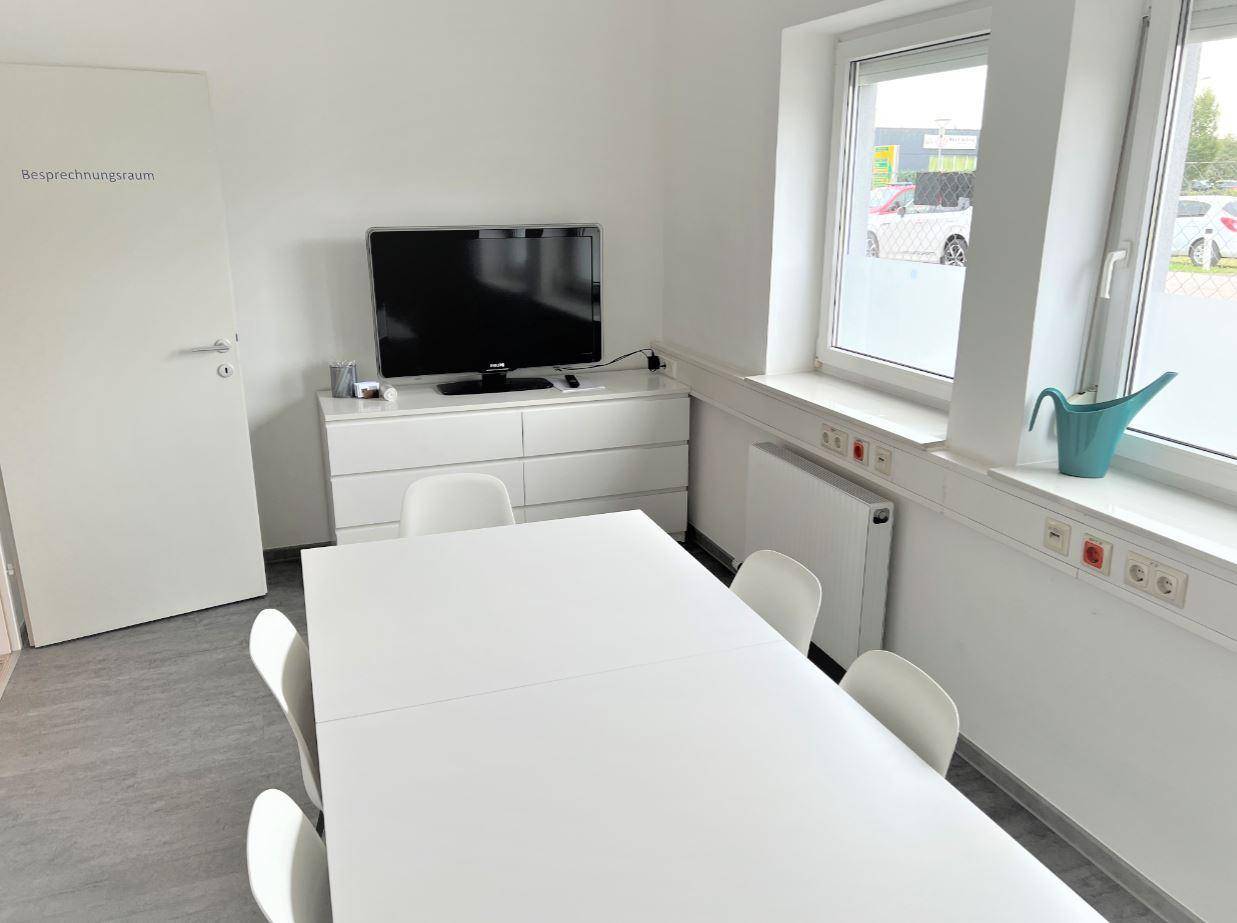 Heller Besprechungsraum mit großem weißen Tisch, modernen Stühlen und einem Fernseher.
