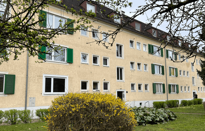 Mehrfamilienhaus mit gelber Fassade, grünen Fensterläden und Bäumen im Vordergrund.