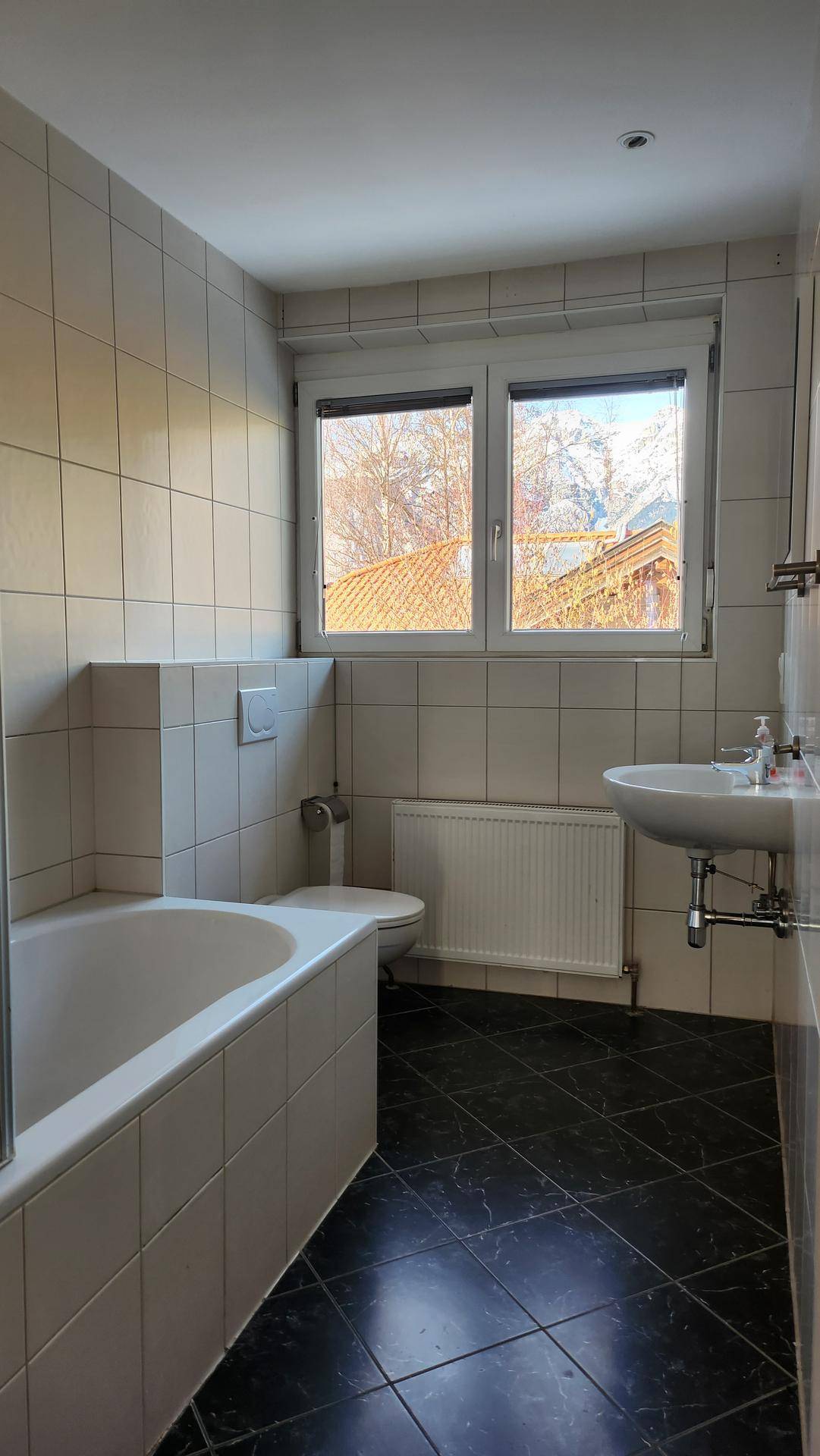 Badezimmer mit Badewanne, WC, Waschbecken und weißen Fliesen, mit Fensterblick.