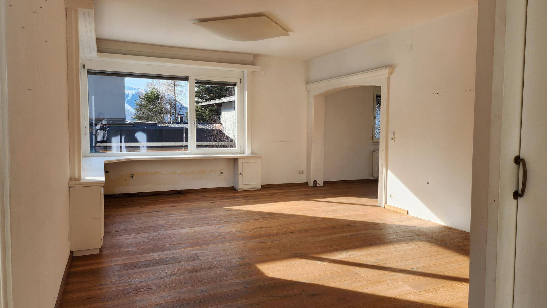 Helles, leeres Wohnzimmer mit Holzboden und großem Fenster, bereit zur individuellen Gestaltung.