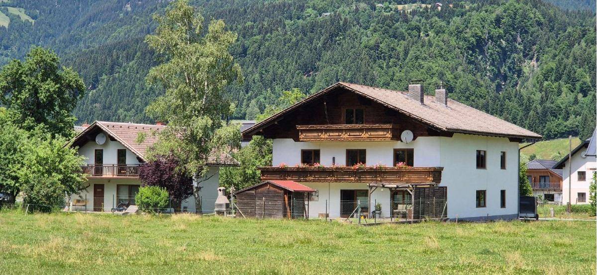 Traditionelles Haus mit weißen Fassaden, Holzbalkonen und einem Satteldach, umgeben von grünen Wiesen und Bergen.