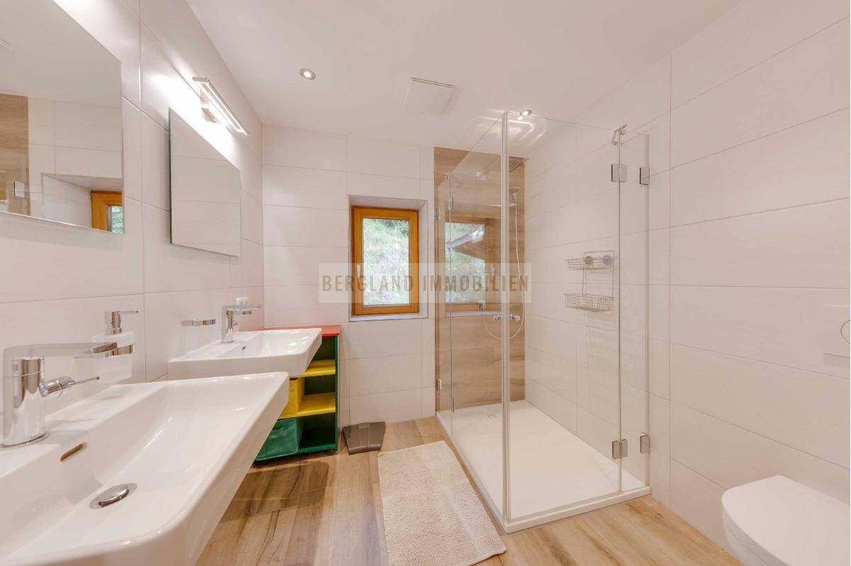 Modernes Badezimmer mit weißen Fliesen, zwei Waschbecken, begehbarer Dusche und Fenster für Tageslicht.