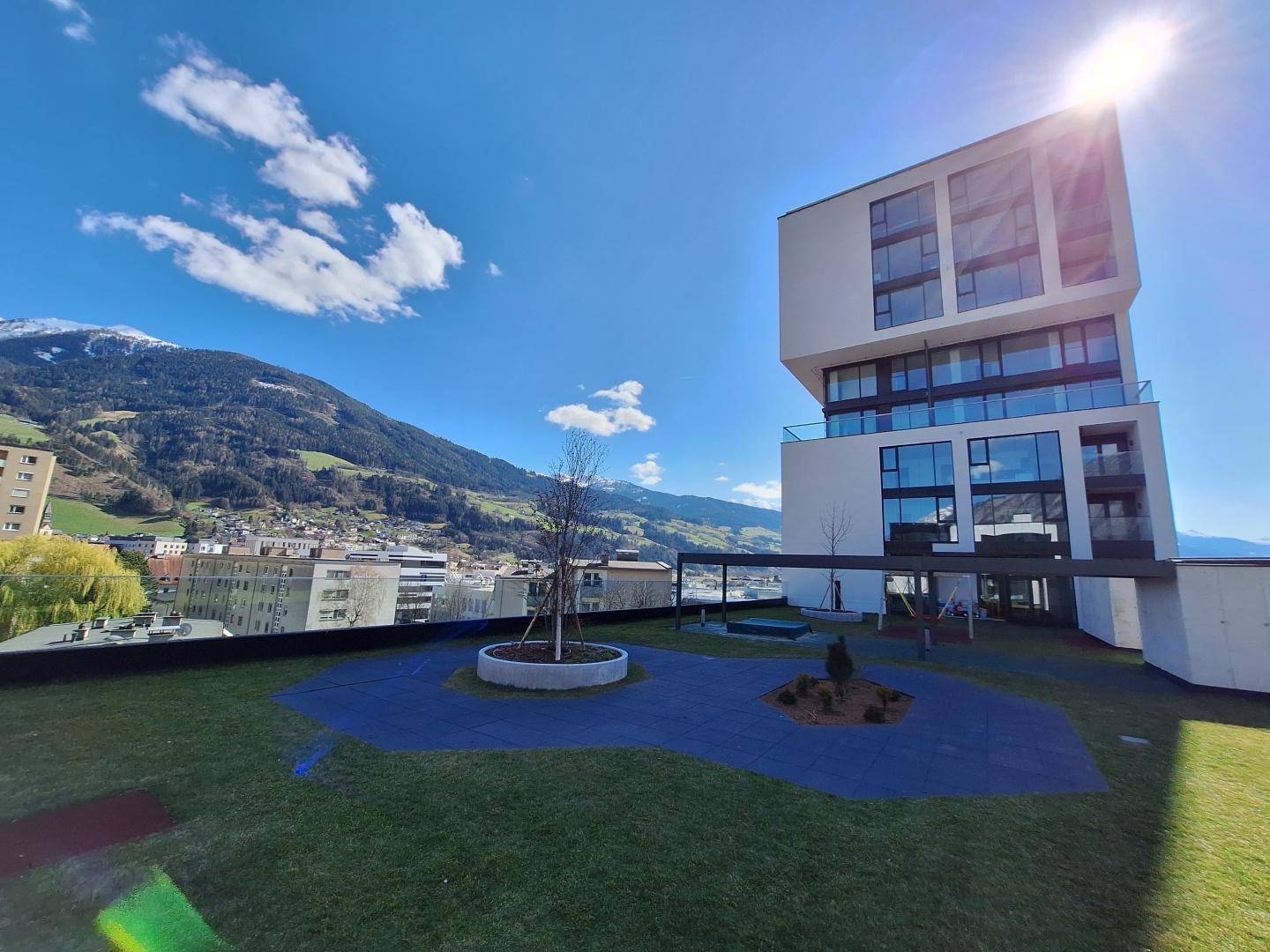 Moderne Wohnanlage mit Gemeinschaftsgarten, Spielplatz und beeindruckendem Bergblick unter blauem Himmel.