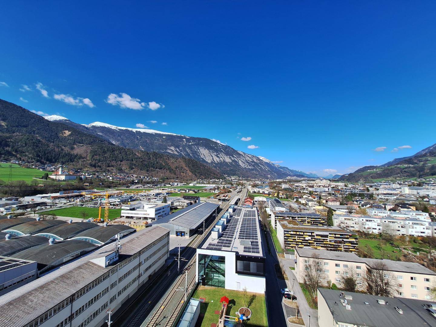 Panoramablick auf eine Stadt im Tal, umgeben von schneebedeckten Bergen unter blauem Himmel.