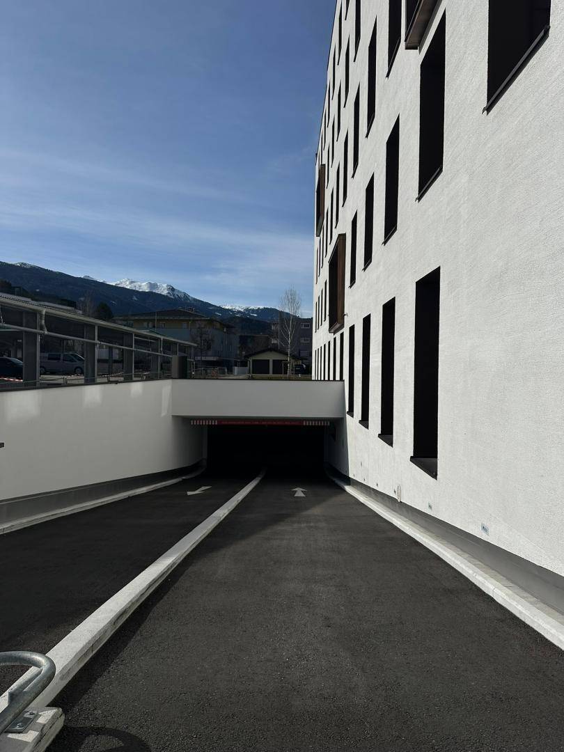 Einfahrt zur Tiefgarage eines modernen Gebäudes mit Rampe und Bergblick im Hintergrund.