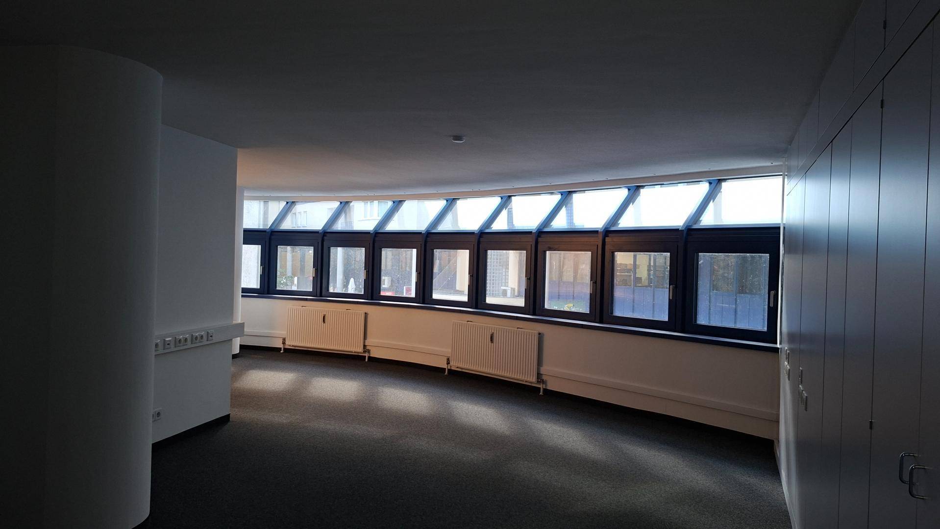 Heller Büroraum mit geschwungener Fensterfront, Heizkörpern und Teppichboden.