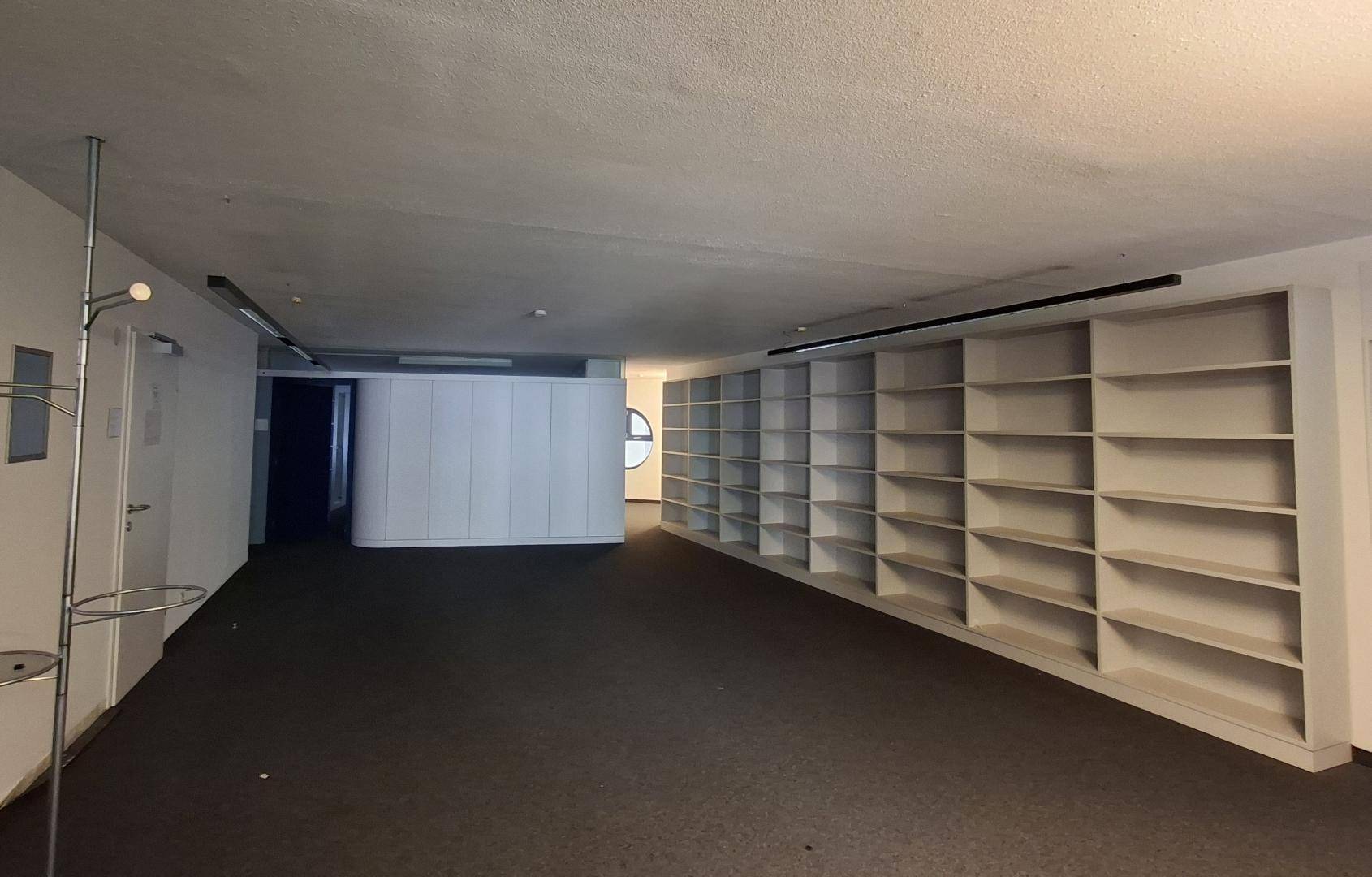 Geräumiger Büroraum mit Teppichboden und einer langen Regalwand für Stauraum.