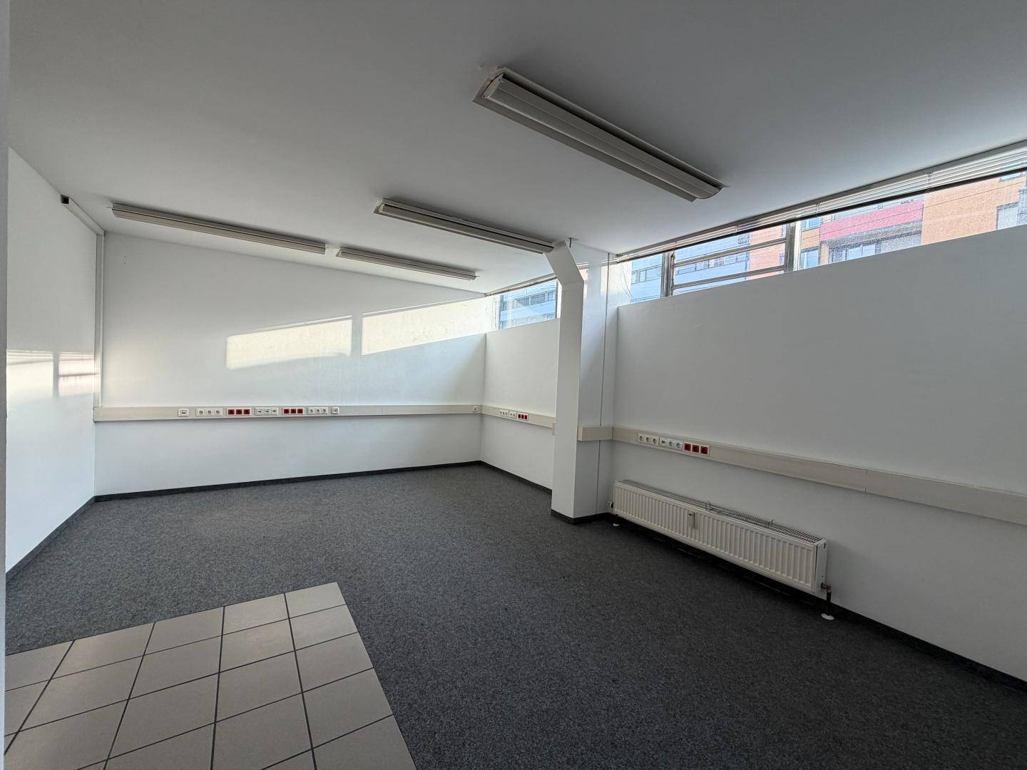 Leerer Büroraum mit grauem Teppichboden, weißen Wänden und Deckenleuchten, ideal für gewerbliche Nutzung.