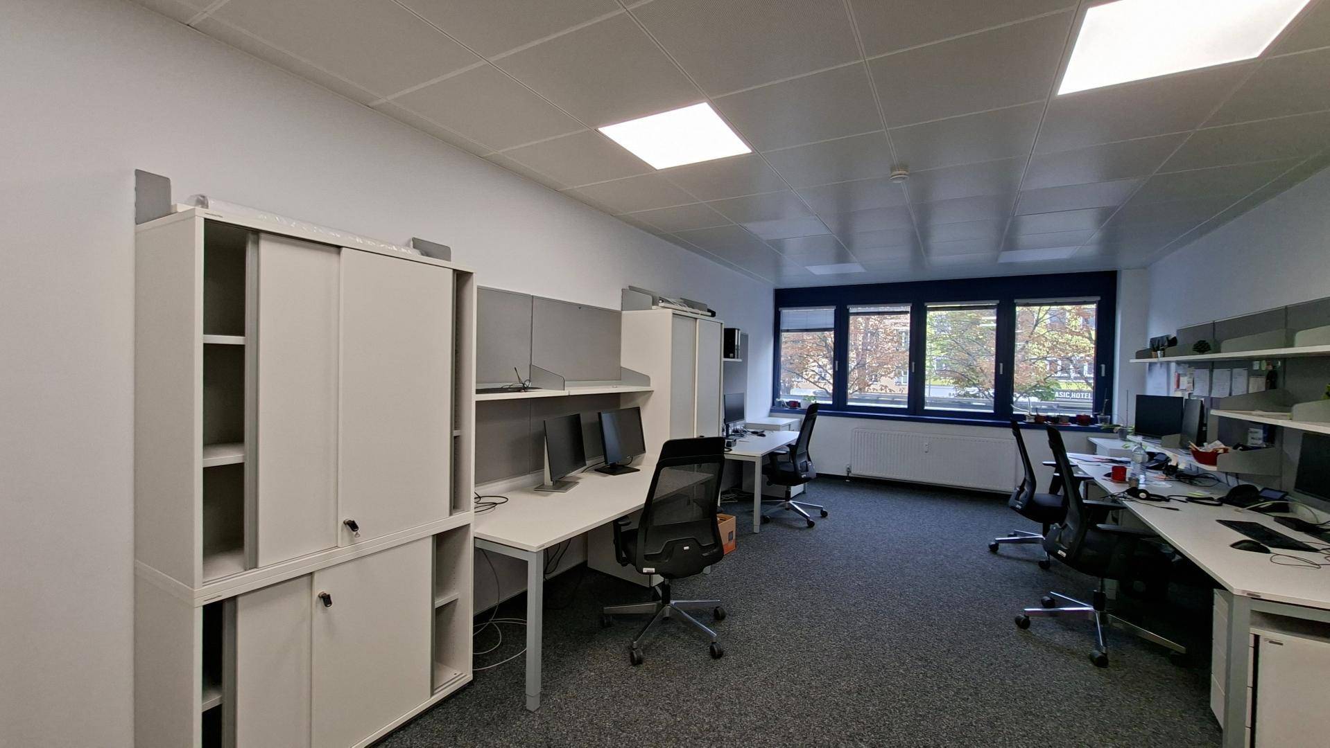 Helles Büro mit mehreren Arbeitsplätzen, modernen Möbeln und großen Fenstern.