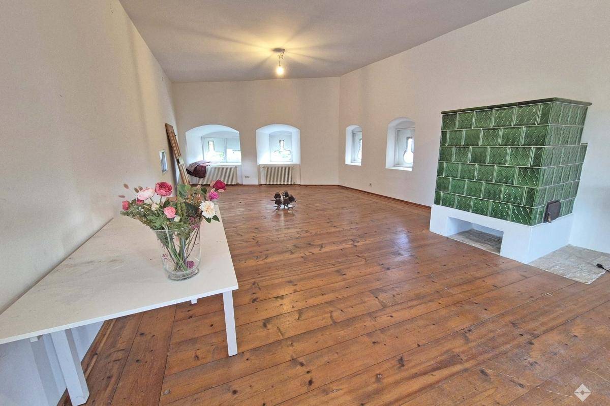 Heller Wohnraum mit Holzdielenboden, Kachelofen und kleinen Fenstern mit tiefen Laibungen.