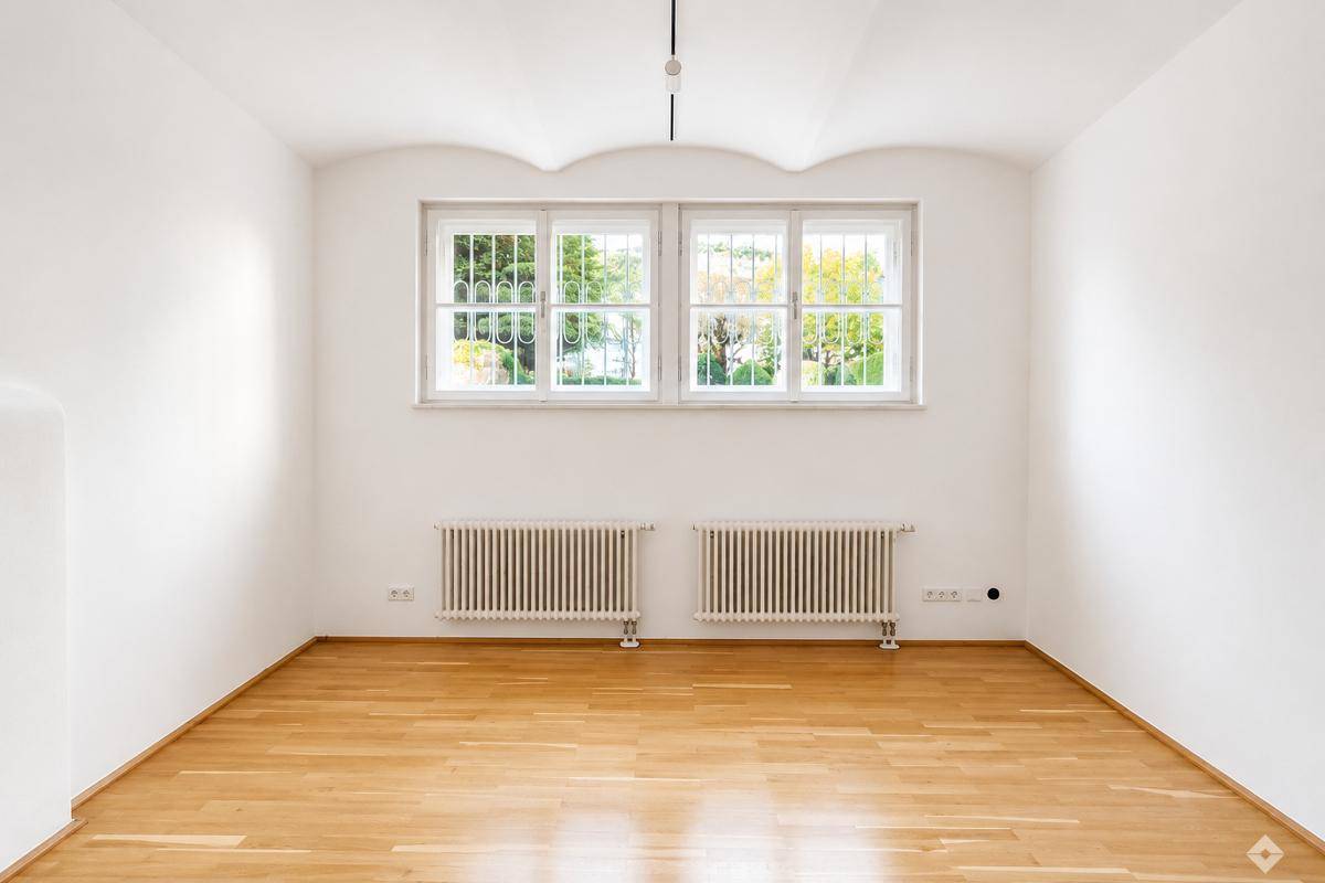 Leeres Zimmer mit Holzboden, weißen Wänden und zwei großen Fenstern, die einen schönen Blick ins Grüne bieten.