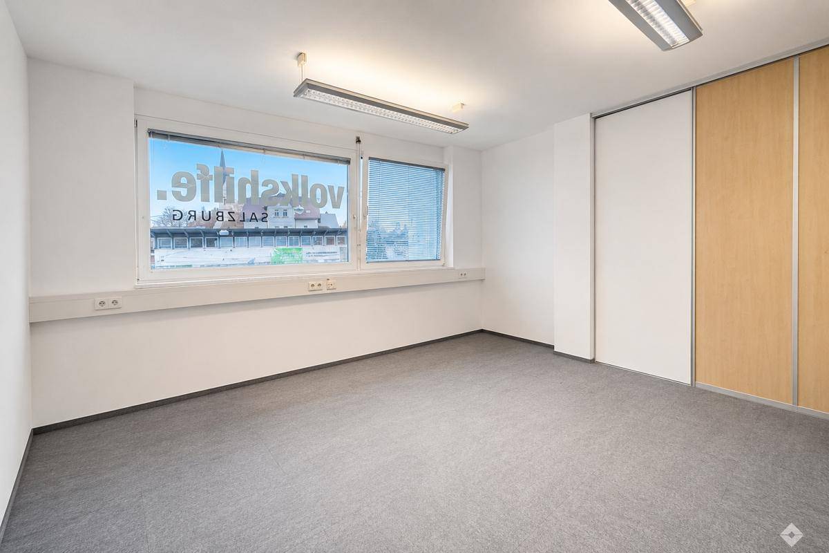 Leeres Büro mit grauem Teppichboden und großen Fenstern, die einen Blick auf die Stadt bieten.