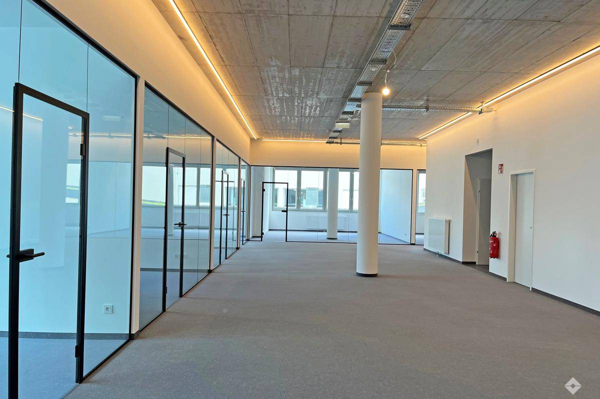 Helles und geräumiges Büro mit Teppichboden, Glaswänden und sichtbarer Deckeninstallation.