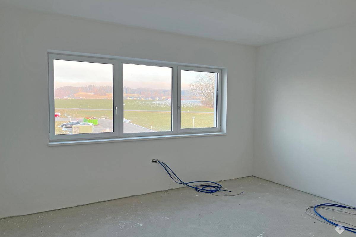 Heller, unfertiger Raum mit weißen Wänden, Betonboden und weitem Blick ins Grüne durch das Fenster.