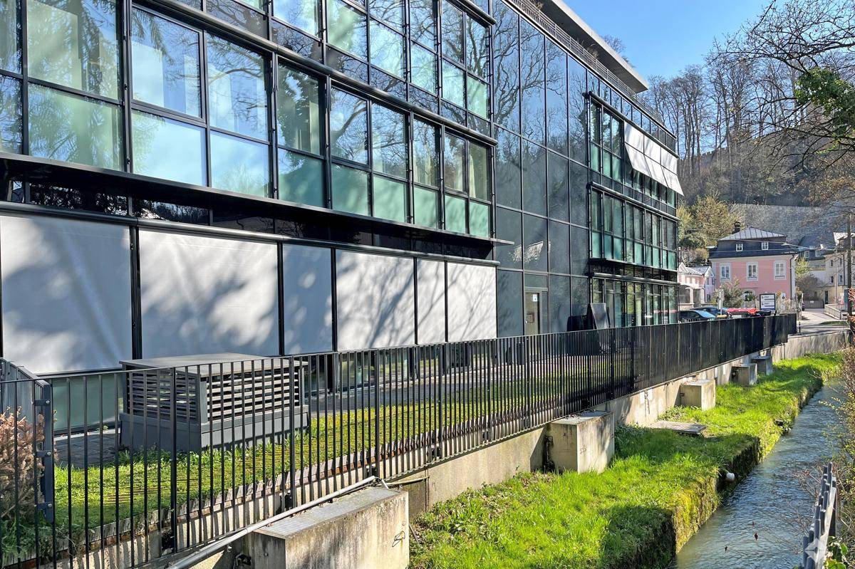 Moderne Glasfassade eines Bürogebäudes entlang eines kleinen Kanals mit grüner Uferböschung und Zaun.