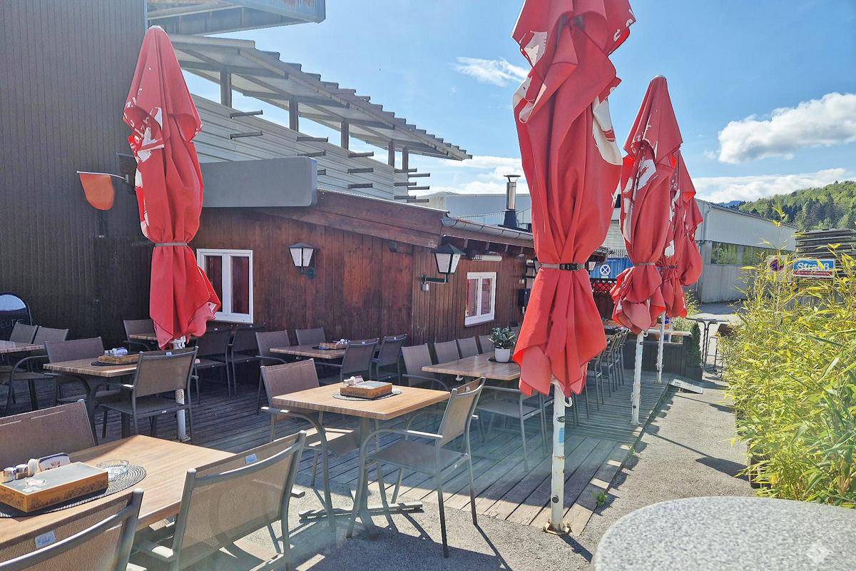 Außenansicht des Holzgebäudes mit einer sonnigen Terrasse und zusammengeklappten roten Sonnenschirmen.